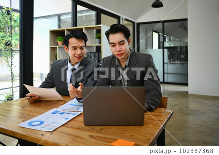 Two young businessmen using laptop discussing project strategy, checking corporate plan at co working office space Two young businessmen using laptop discussing project strategy, checking corporate plan at co working office space 102337368
