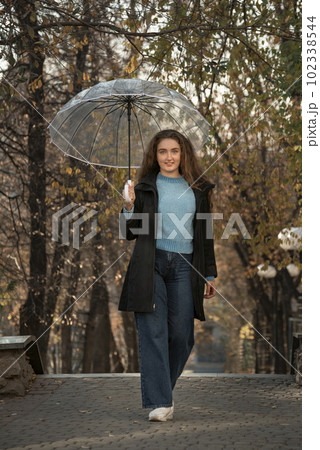 Pretty young woman with lush hair standing with transparent umbrella in autumn park. Photo of woman in park Pretty young woman with lush hair standing with transparent umbrella in autumn park. Photo of woman in park 102338544
