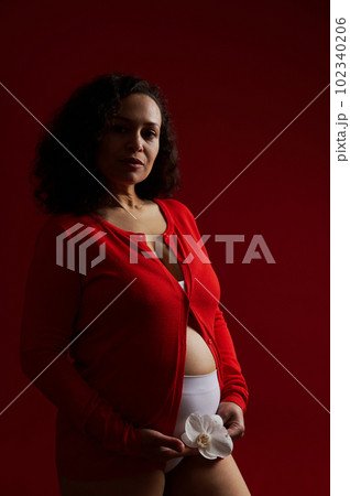 Confident portrait of ethnic pregnant woman, authentic expectant mother wearing red unbuttoned shirt, holding white orchid flower, looking at camera, posing bare belly on dark red isolated background Confident portrait of ethnic pregnant woman, authentic expectant mother wearing red unbuttoned shirt, holding white orchid flower, looking at camera, posing bare belly on dark red isolated background 102340206