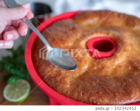 Freshly-baked Savarin in red silicone cake mold is poured by syrup. Rum-soaked circular cake, close up Freshly-baked Savarin in red silicone cake mold is poured by syrup. Rum-soaked circular cake, close up 102342452