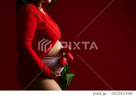 Closeup pregnant woman dressed in red shirt and white underwear, holding bunch of red tulips, touching her belly on isolated background. Copy ad space. Happy Mother's Day concept. pregnancy. Maternity 102361306