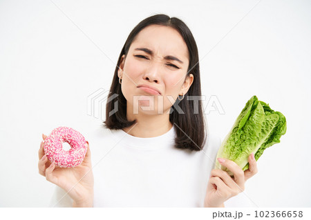 Healthy clean detox eating concept. Vegetarian, vegan, raw concept. Sad asian girl on diet holds doughnut and vegetable, wants to taste donnut, white background Healthy clean detox eating concept. Vegetarian, vegan, raw concept. Sad asian girl on diet holds doughnut and vegetable, wants to taste donnut, white background 102366658