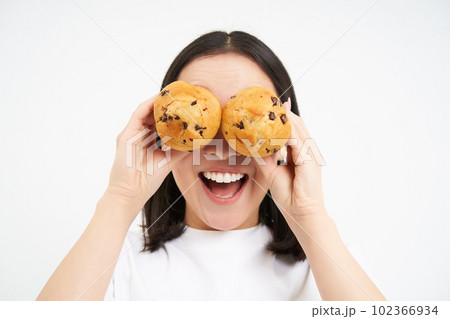 Close up portrait of funny asian girl, holds cupcakes near face, makes happy faces, white studio background Close up portrait of funny asian girl, holds cupcakes near face, makes happy faces, white studio background 102366934