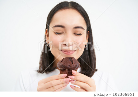 Pastry, sweets and bakery. Close up of smiling asian woman, biting cupcake with joy, standing isolated on white background Pastry, sweets and bakery. Close up of smiling asian woman, biting cupcake with joy, standing isolated on white background 102366951