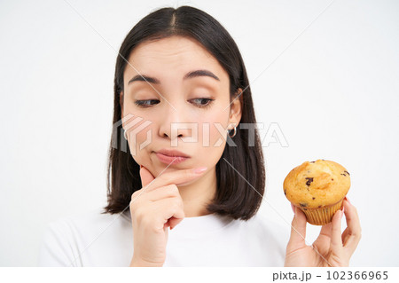 Close up portrait of asian woman with pensive smiling face, holds cupcake, thinks to eat pastry, white background Close up portrait of asian woman with pensive smiling face, holds cupcake, thinks to eat pastry, white background 102366965