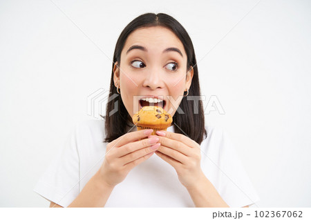 Close up of happy korean woman eating cupcake, delicious dessert, smiling from pleasure, white background Close up of happy korean woman eating cupcake, delicious dessert, smiling from pleasure, white background 102367062