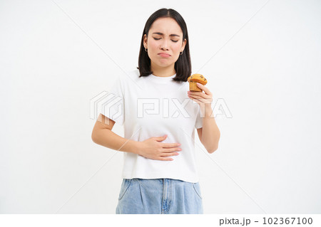 Portrait of korean woman has stomach ache, touching belly, holding cupcake, cant eat pastry, white studio background 102367100