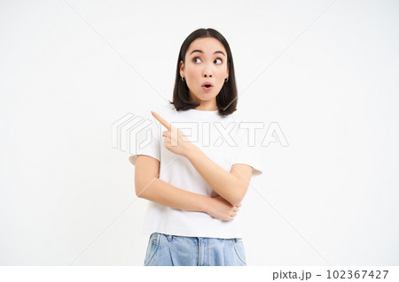 Surprised korean girl, points left and looks with amazed face expression, stands in tshirt isolated on white background 102367427
