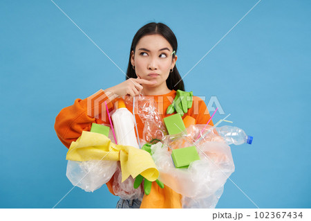 Asian woman holds plastic waste and makes thinking face, ponders how to recycle bottles and household items, blue background Asian woman holds plastic waste and makes thinking face, ponders how to recycle bottles and household items, blue background 102367434