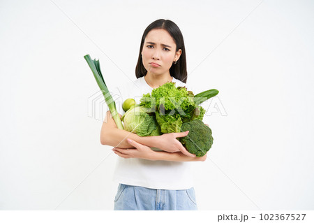 Image of sad asian girl, holding vegetables and sulking, being on diet, white background Image of sad asian girl, holding vegetables and sulking, being on diet, white background 102367527