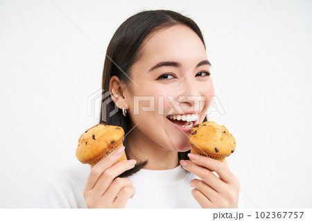 Image of happy korean girl who loves pastry, smiling and eating cupcakes, isolated on white background 102367757