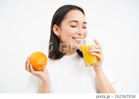 Close up portrait of asian woman, drinking glass of orange juice with delight and happy smile, white studio background 102367836