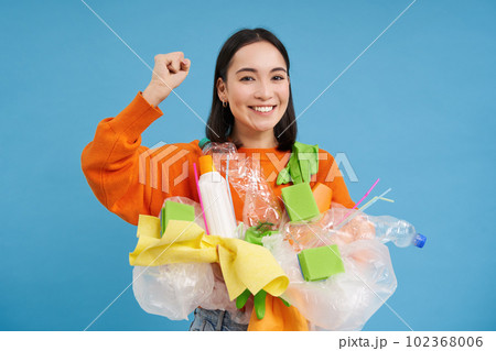 Enthusiastic asian woman holding non sorted plastic garbage, recycling materials with happy face, standing over blue background Enthusiastic asian woman holding non sorted plastic garbage, recycling materials with happy face, standing over blue background 102368006