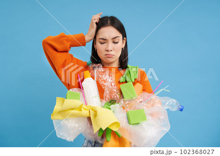 Confused Korean woman tries to go green, recycle plastic household items, holding bottles and gloves, does recycling, blue background Confused Korean woman tries to go green, recycle plastic household items, holding bottles and gloves, does recycling, blue background 102368027