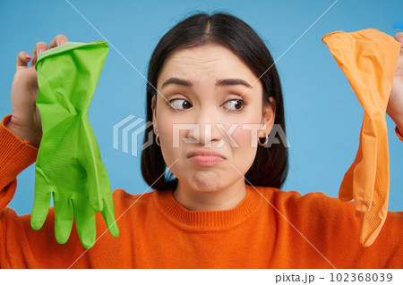 Close up portrait of reluctant young woman, showing two different latex gloves for cleaning, dislikes washing and cleaning, blue background Close up portrait of reluctant young woman, showing two different latex gloves for cleaning, dislikes washing and cleaning, blue background 102368039