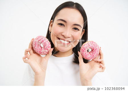 Close up portrait of cute korean woman, smiling while eating pink glazed doughnuts, likes tasty donnuts, white background Close up portrait of cute korean woman, smiling while eating pink glazed doughnuts, likes tasty donnuts, white background 102368094