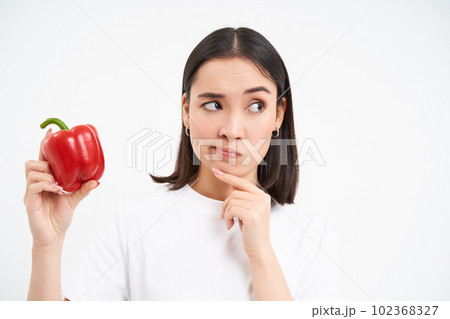 Close up portrait of asian woman, holding red sweet pepper and thinking, stands with pensive face, white background Close up portrait of asian woman, holding red sweet pepper and thinking, stands with pensive face, white background 102368327