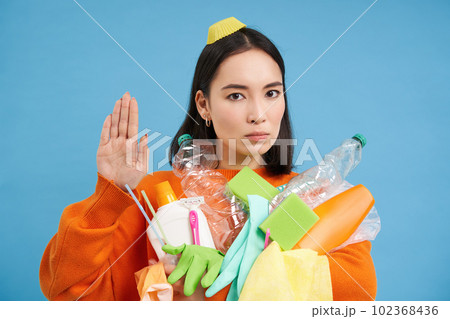 Portrait of serious asian woman shows stop sign, holds plastic for recycling, stop throwing waste into general bin, blue background Portrait of serious asian woman shows stop sign, holds plastic for recycling, stop throwing waste into general bin, blue background 102368436