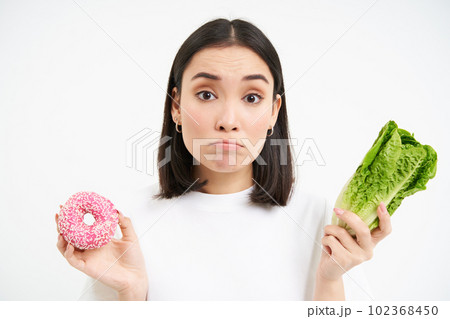 Healthy lifestyle concept. Portrait of sad complicated asian woman, shows doughnut and cabbage, decides between vegetables and diet or delicious junk food, white background Healthy lifestyle concept. Portrait of sad complicated asian woman, shows doughnut and cabbage, decides between vegetables and diet or delicious junk food, white background 102368450