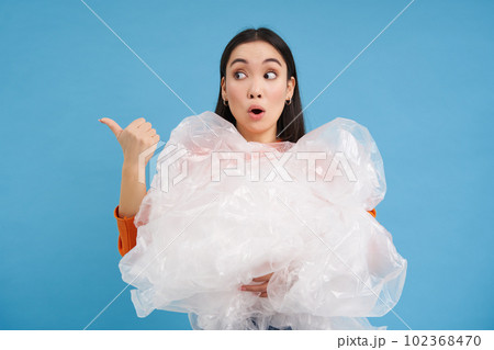 Woman with surprised face, holding plastic recycling waste and pointing left with amazed emotion, blue background 102368470