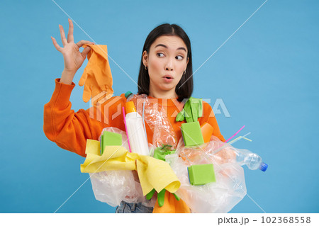 Young asian woman holds garbage, plastic bottles and waste, looks surprised at dirty latex glove, blue background Young asian woman holds garbage, plastic bottles and waste, looks surprised at dirty latex glove, blue background 102368558