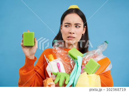 Portrait of asian woman looking disgusted at dirty sponge, holding recycable empty bottles, sorting garbage, blue background Portrait of asian woman looking disgusted at dirty sponge, holding recycable empty bottles, sorting garbage, blue background 102368719