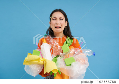 Crying asian female holding plastic waste, garbage in hands and looking upset, has to recycle, blue background Crying asian female holding plastic waste, garbage in hands and looking upset, has to recycle, blue background 102368780