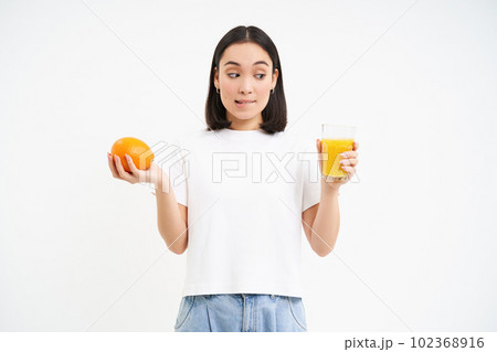 Cute korean girl, makes choice between orange fuit and juice, looks both sides, white background 102368916