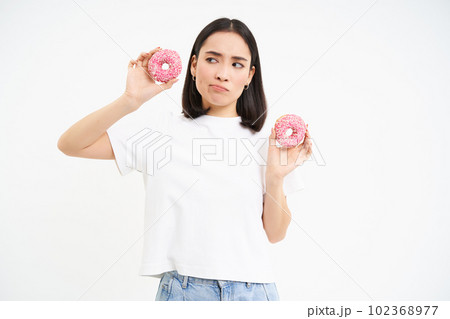 Thoughtful korean woman, holding two glazed doughnuts, thinking, being on diet, white studio background 102368977
