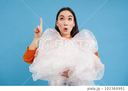 Surprised korean woman with plastic waste, points finger up and looks on top, blue background Surprised korean woman with plastic waste, points finger up and looks on top, blue background 102368991