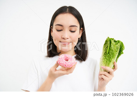 Healthy clean detox eating concept. Vegetarian, vegan, raw concept. Smiling korean girl eats delicious doughnut, holds cabbage, enjoys junk food, white background 102369026