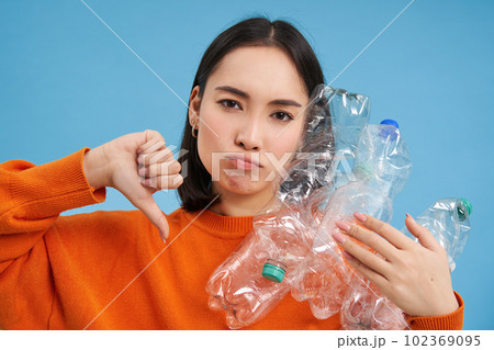 Portrait of woman with upset face, shows thumb down and plastic bottles, disappointed with lack of recycling, blue background 102369095
