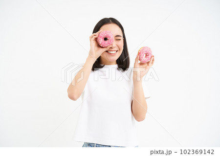 Portrait of happy korean woman, makes glasses from two tasty glazed doughnuts, smiling and having fun, white background 102369142