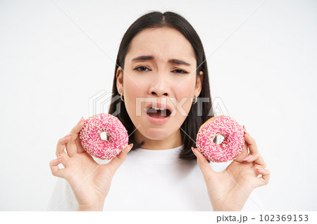 Close up portrait of sad asian woman, upset being on diet, showing two glazed pink doughnuts, tempted to eat junk food, white background Close up portrait of sad asian woman, upset being on diet, showing two glazed pink doughnuts, tempted to eat junk food, white background 102369153