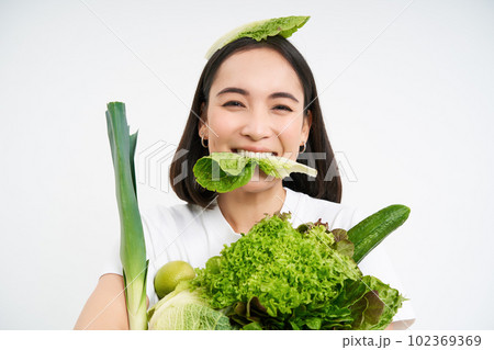 Portrait of smiling korean woman eats lettuce leaf, holding green vegetables, eating nutritious food, white background 102369369