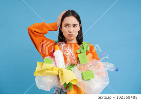 Confused girl with plastic garbage in hands, scratches head, looks puzzled with recycling system, blue background 102369488