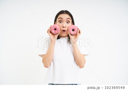 Beautiful young woman eating delicious glazed dougnuts, holding two pink donuts and smiling, white background Beautiful young woman eating delicious glazed dougnuts, holding two pink donuts and smiling, white background 102369549