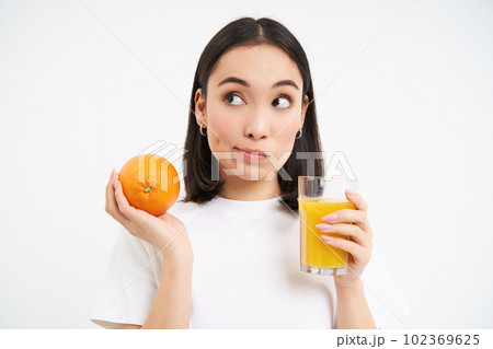 Close up portrait of asian woman with orange and glass of juice, thinking, making choice, standing over white background 102369625