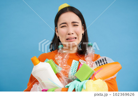Portrait of crying sad girl with plastic trash, collects recycling waste, looks miserable, blue background 102369626