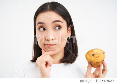 Close up portrait of korean woman with one cupcake, thinking and smiling, white studio background Close up portrait of korean woman with one cupcake, thinking and smiling, white studio background 102369627