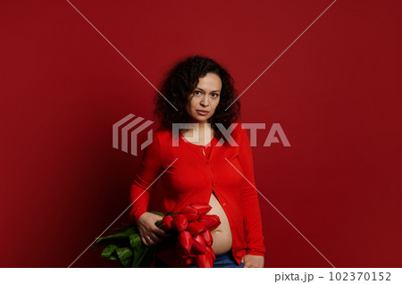 Multi ethnic curly haired pregnant woman looking confidently at camera, holding a bouquet of red tulips on isolated color background, dressed in bright red shirt and blue jeans. Pregnancy fashion Multi ethnic curly haired pregnant woman looking confidently at camera, holding a bouquet of red tulips on isolated color background, dressed in bright red shirt and blue jeans. Pregnancy fashion 102370152
