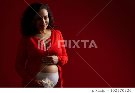 Beautiful multi ethnic pregnant woman, expectant mother holding white orchid flower, smiling, gently caressing her belly, enjoying happy moments of pregnancy and maternity, isolated on red background Beautiful multi ethnic pregnant woman, expectant mother holding white orchid flower, smiling, gently caressing her belly, enjoying happy moments of pregnancy and maternity, isolated on red background 102370226