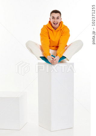 young man in orange sweatshirt sitting on white cube 102371255
