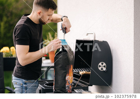 Man opens a package of charcoal for grilling. Preparing for barbecue on backyard Man opens a package of charcoal for grilling. Preparing for barbecue on backyard 102375978