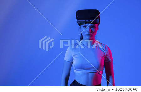 Young brunette woman wearing virtual reality device, vr glasses and playing a game with a friends online in neon light. Young brunette woman wearing virtual reality device, vr glasses and playing a game with a friends online in neon light. 102378040