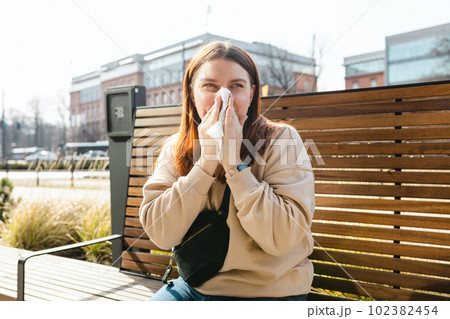 Unhappy caucasian millennial red-haired woman sneezing in napkin and siting in park bench, Young women suffering from allergy outdoors 102382454