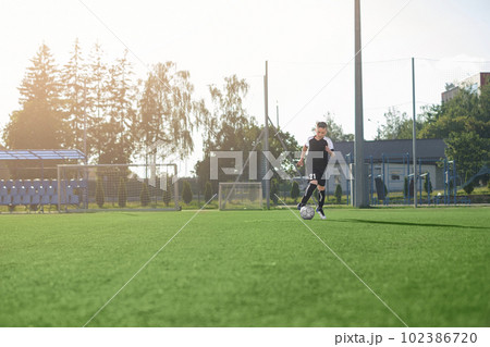 A teenage boy in a black sports uniform with the number twenty-one on his shorts is training to play football at the stadium. Sports events and junior soccer matches 102386720