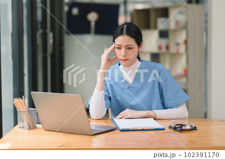 A concentrated female doctor or nurse is working online with a laptop while sitting at a desk in a hospital consultation room. medicine, technology and healthcare concept A concentrated female doctor or nurse is working online with a laptop while sitting at a desk in a hospital consultation room. medicine, technology and healthcare concept 102391170
