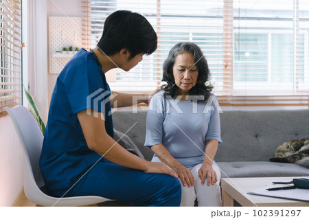 A young caregiver is consulting with a 60-year-old Asian elderly woman, providing encouragement and taking a medical history. The two are shown holding hands and exchanging words of encouragement. 102391297