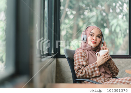 A confident,beautiful, professional, millennial Muslim Asian businesswoman wearing a brown hijab is relaxing in her office and having coffee. A confident,beautiful, professional, millennial Muslim Asian businesswoman wearing a brown hijab is relaxing in her office and having coffee. 102391376